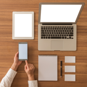 Overhead view of desk with laptop, iPad, notebook, pen, flashcards, and white hands holding phone.