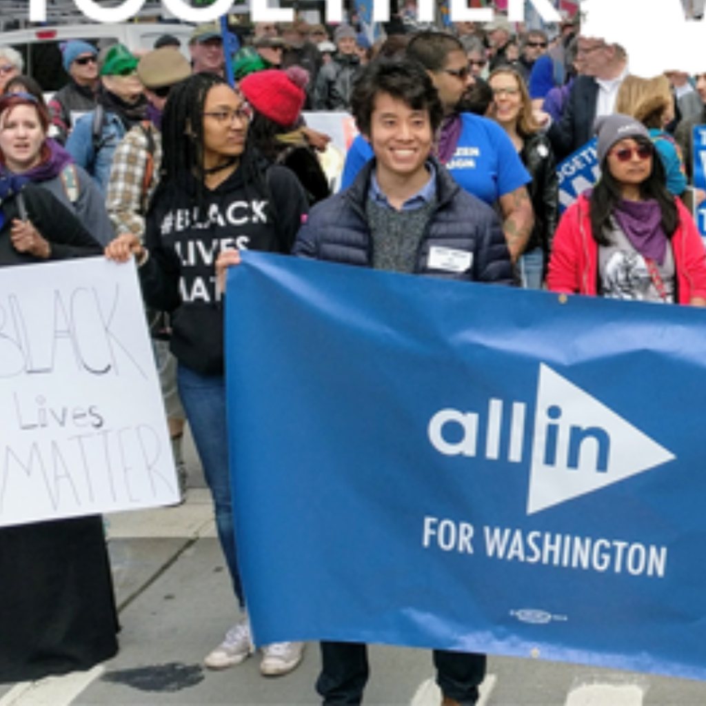 A crowd of diverse people participates in a march, holding banners that say "Black Lives Matter" and "All In for Washington."