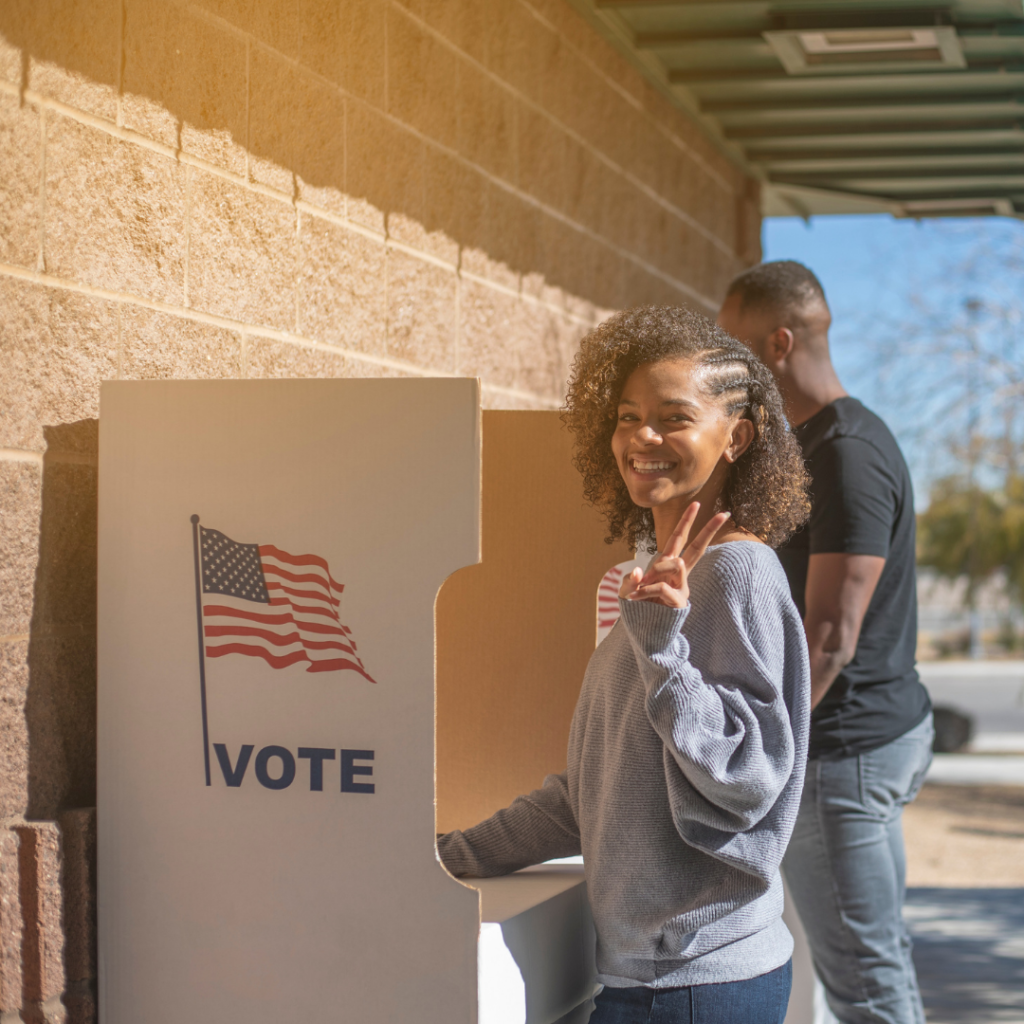 Young African American woman making peace sign with fingers while smiling at camera, standing at a voting booth with an American flag and text that says "VOTE"